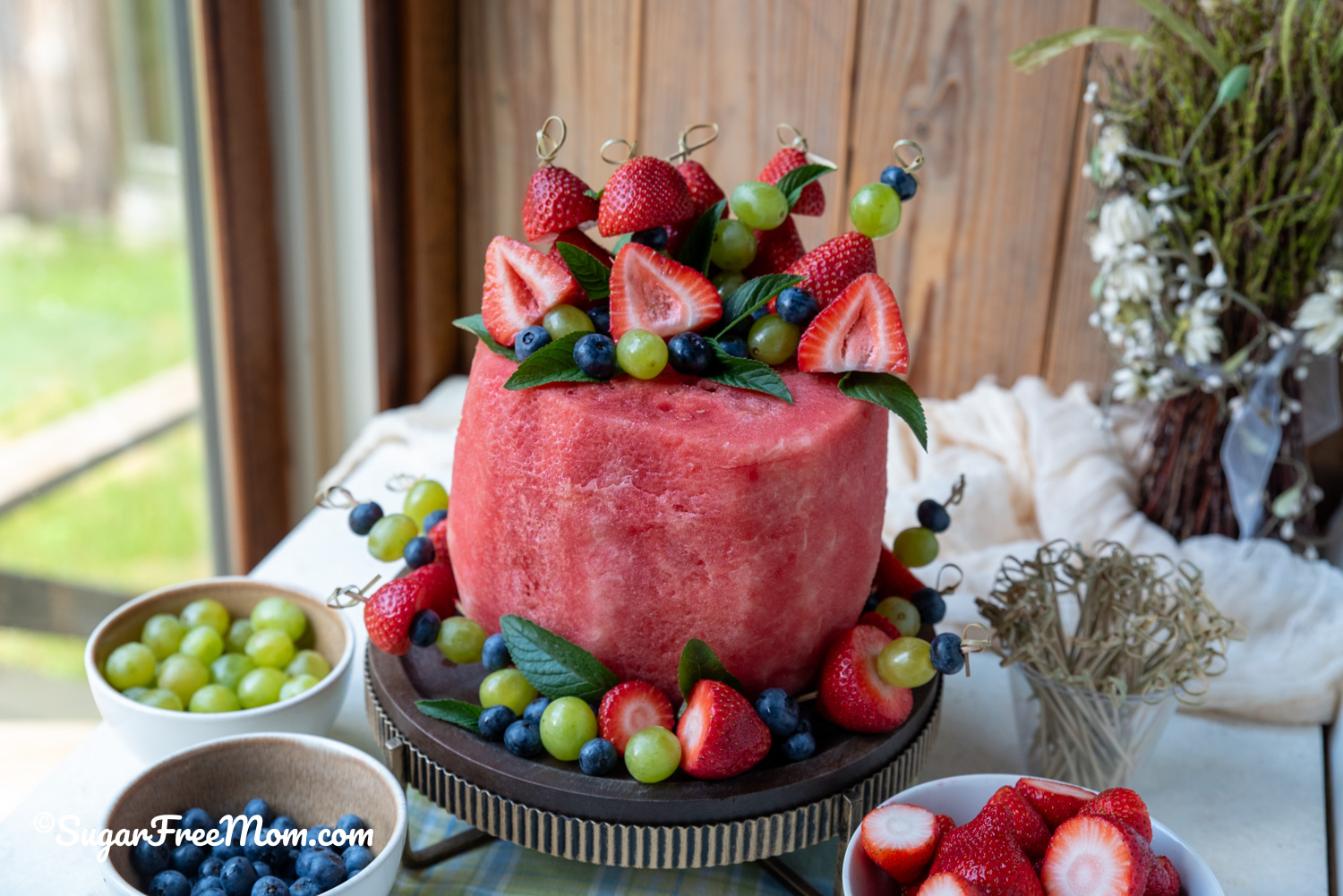 horizontal pic of a fresh fruit watermelon cake on a stand with fruit skewers