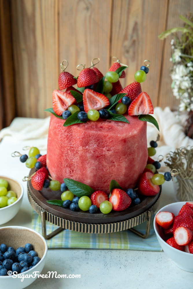 vertical pic of watermelon cake on a stand with fruit skewers