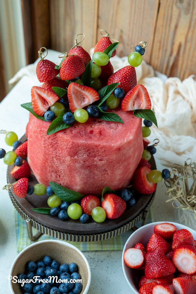 vertical pic of a fresh fruit watermelon cake on a stand with fruit skewers